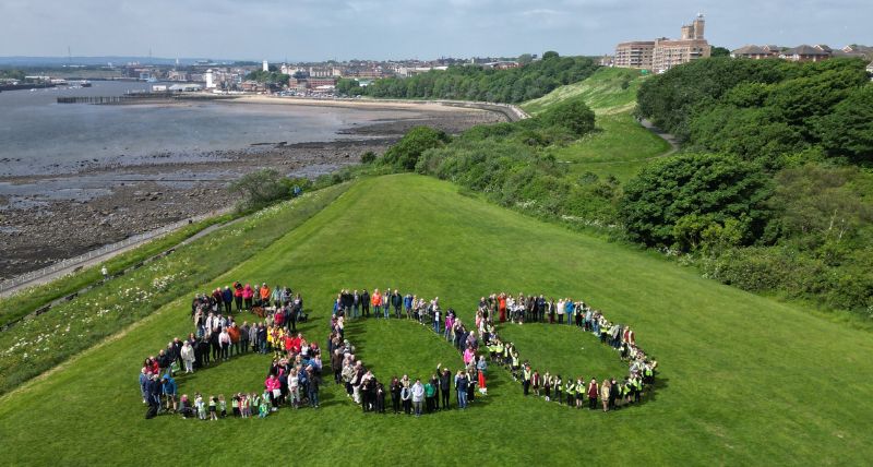 Dozens of people standing in the shape of an 800 on a grassed area with the river behind